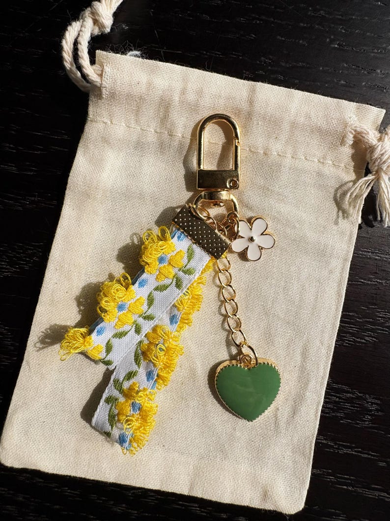 May include: A floral keychain with a gold clasp and chain. The keychain features a white ribbon with yellow and blue flower embroidery, yellow fringe, a white flower charm, and a green heart charm. The keychain rests on a beige drawstring bag.