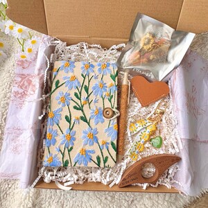 May include: A gift box filled with a floral embroidered journal, pen, heart-shaped leather tag, keychain, tea bag, and a wooden page holder. The journal features blue flowers on a beige background.