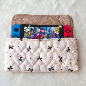 May include: A pink quilted case with a heart pattern and black bow accents, designed to hold a Nintendo Switch. The case has a floral patterned top flap and a small label that reads "Handmade with love."
