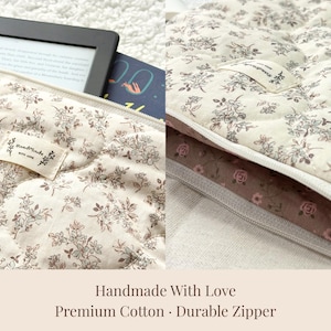 May include: Cream-colored quilted fabric case with a floral pattern, holding an e-reader and a book. The case has a durable zipper and a small tag that says "handmade with love". The bottom of the case is a dark brown fabric with a rose pattern.