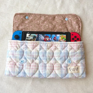 May include: A pastel pink and blue plaid quilted case for a gaming console, with a heart pattern on the front. The interior is brown with a floral print. The case is open, showing a console with blue and red controllers. Fastened with light blue buttons.
