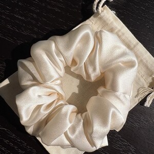 May include: A cream-colored satin scrunchie rests on a small, beige drawstring bag. The scrunchie is gathered and voluminous, with a soft, lustrous sheen. The bag is made of a natural-colored fabric. The items are on a dark wooden surface.