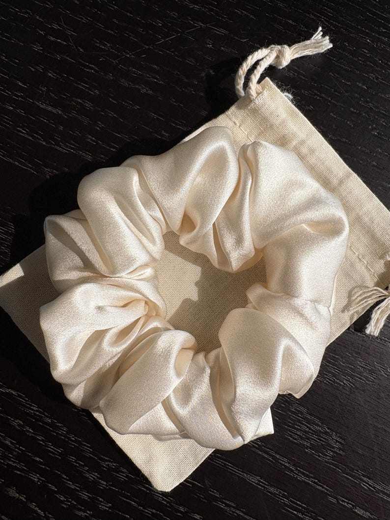 May include: A cream-colored satin scrunchie rests on a small, beige drawstring bag. The scrunchie is gathered and voluminous, with a soft, lustrous sheen. The bag is made of a natural-colored fabric. The items are on a dark wooden surface.