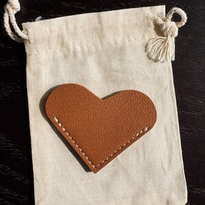 May include: A small, off-white drawstring bag with a brown leather heart-shaped patch. The heart is stitched with white thread. The bag has a knotted drawstring closure and a tassel.