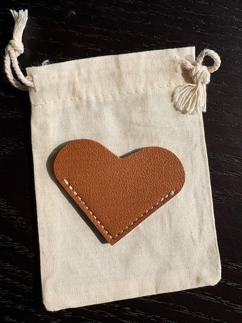 May include: A small, off-white drawstring bag with a brown leather heart-shaped patch. The heart is stitched with white thread. The bag has a knotted drawstring closure and a tassel.