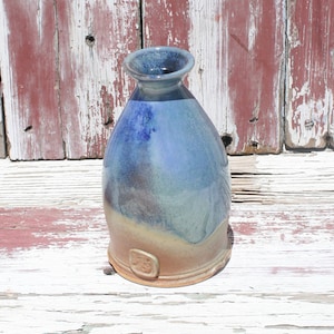 May include: A small, blue and brown ceramic vase with a rounded body and a narrow neck. The vase is sitting on a weathered wooden surface.