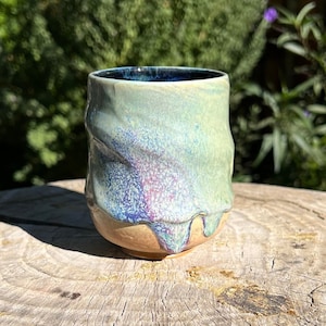 Könnte beinhalten: Eine kleine, grüne, blaue und violette Keramiktasse mit einer strukturierten, geschwungenen Glasur. Die Tasse steht auf einem Baumstumpf.