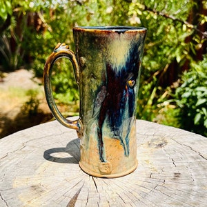 Puede incluir: Una taza de cerámica hecha a mano con un esmalte azul, verde y marrón. La taza tiene un diseño único y texturizado y una asa.