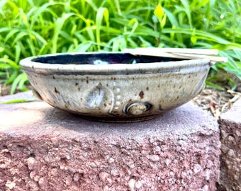 May include: A handmade ceramic bowl with a speckled, tan exterior and a dark interior. The bowl has a rounded shape with decorative indentations and is sitting on a red brick surface. The background features green foliage.