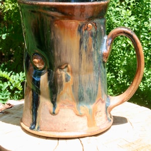 May include: A handmade ceramic mug with a brown, blue, and green glaze. The mug has a unique, textured surface and a large handle.