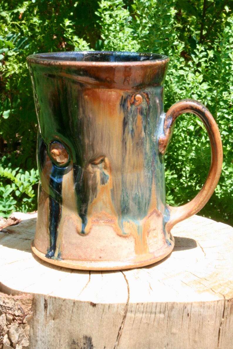 May include: A handmade ceramic mug with a brown, blue, and green glaze. The mug has a unique, textured surface and a large handle.