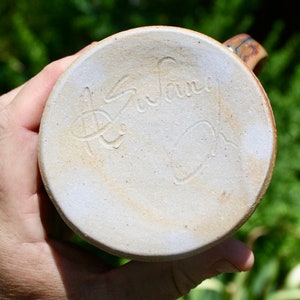 May include: The bottom of a ceramic mug with a light brown glaze. The mug is signed "Susan Oh" in a cursive script.