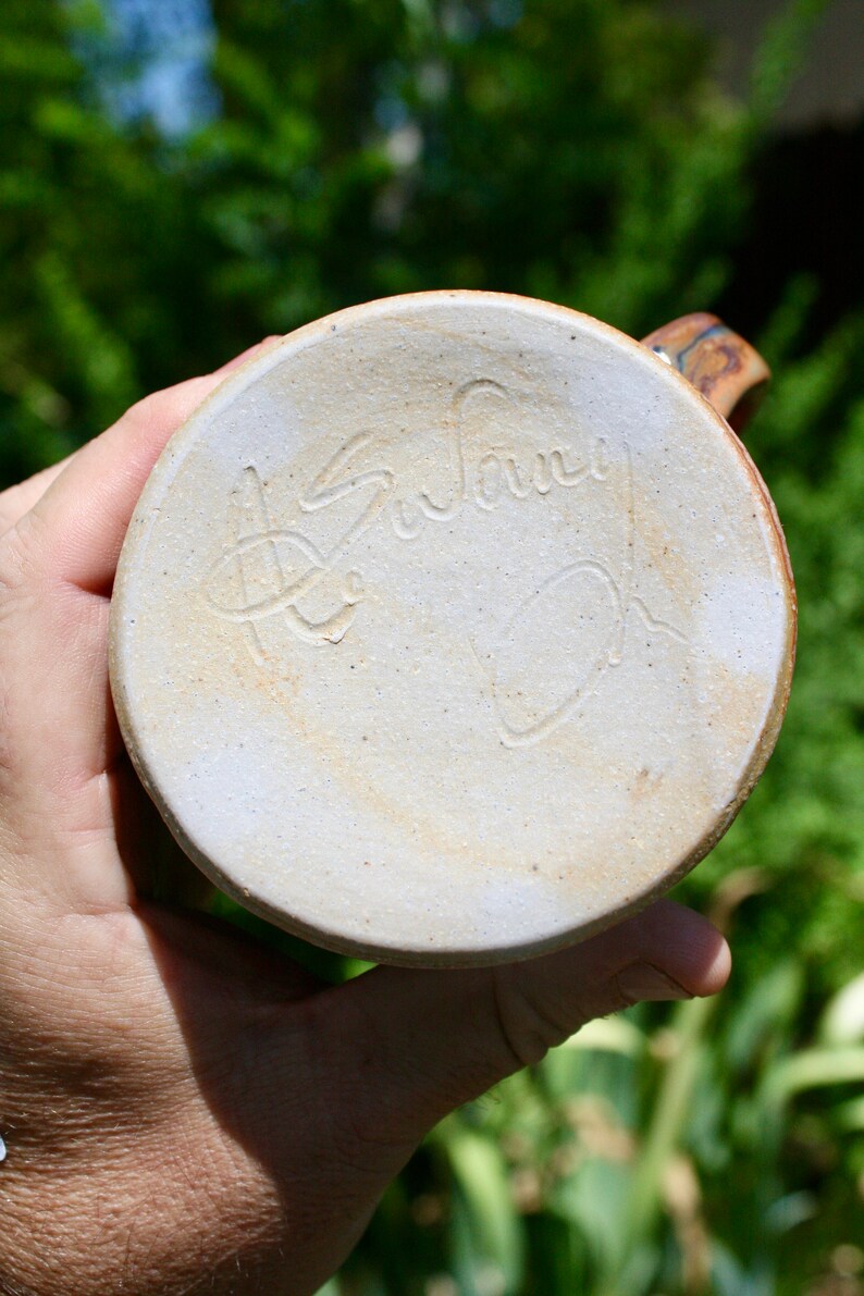 May include: The bottom of a ceramic mug with a light brown glaze. The mug is signed "Susan Oh" in a cursive script.