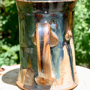 May include: A handmade ceramic mug with a dark green, brown, and black glaze. The mug has a unique, sculpted handle and a rounded body.