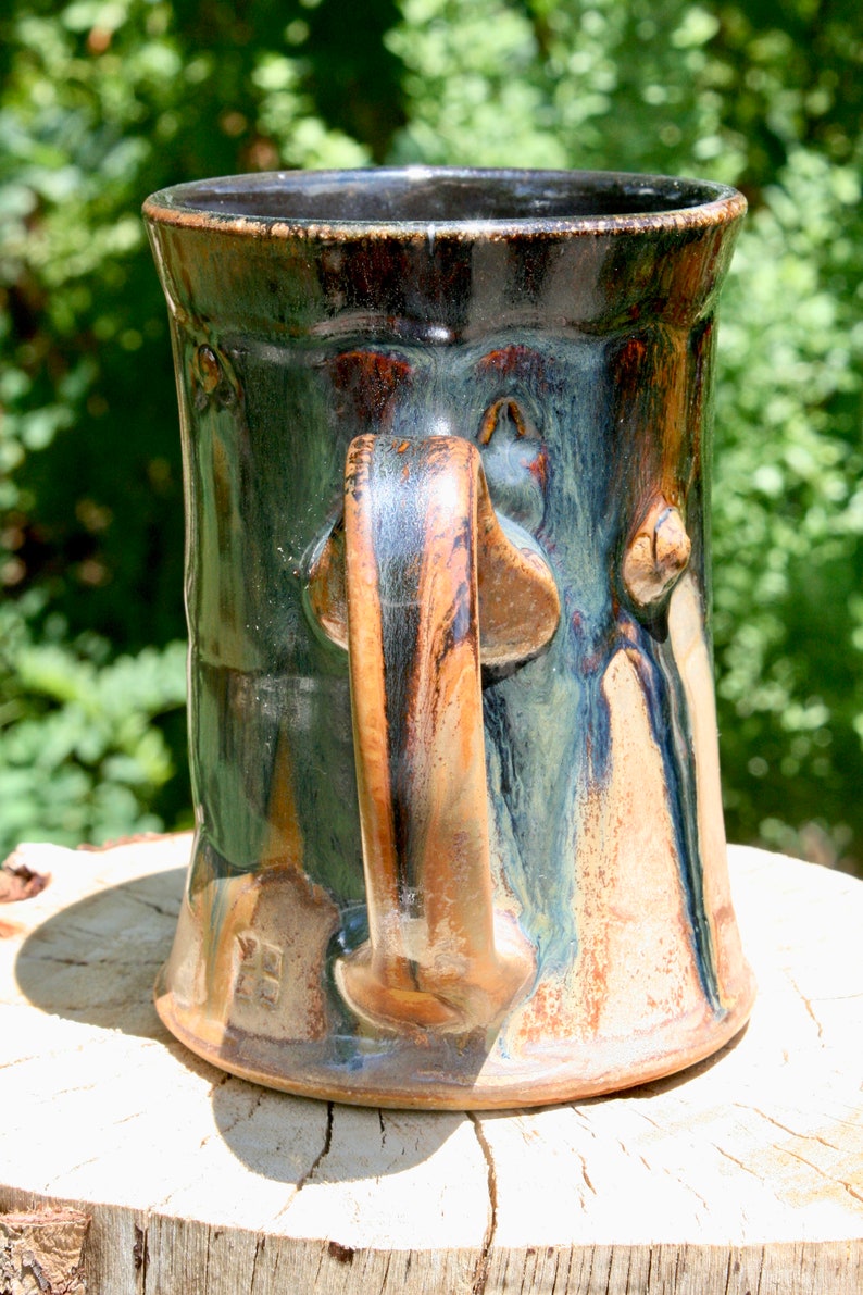 May include: A handmade ceramic mug with a dark green, brown, and black glaze. The mug has a unique, sculpted handle and a rounded body.