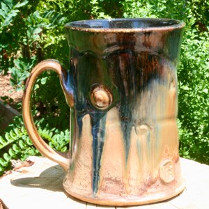 May include: A large, handmade ceramic mug with a brown, green, and blue glaze. The mug has a unique, textured surface that resembles tree bark. The mug has a large handle and a wide opening.