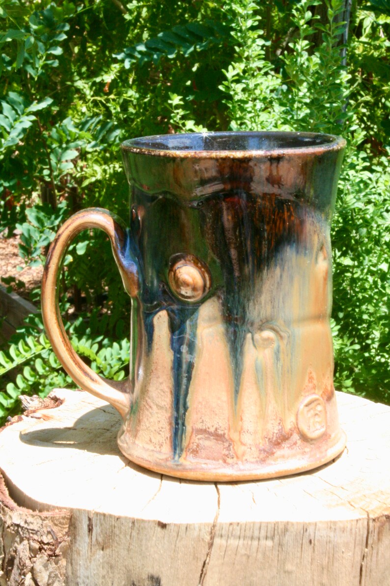 May include: A large, handmade ceramic mug with a brown, green, and blue glaze. The mug has a unique, textured surface that resembles tree bark. The mug has a large handle and a wide opening.