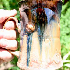 May include: A handmade ceramic mug with a brown, blue, and white glaze. The mug has a unique, textured surface and a rounded handle.