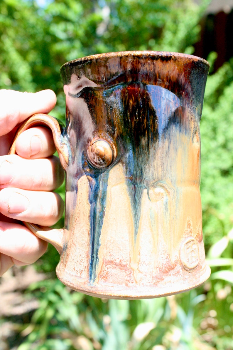 May include: A handmade ceramic mug with a brown, blue, and white glaze. The mug has a unique, textured surface and a rounded handle.