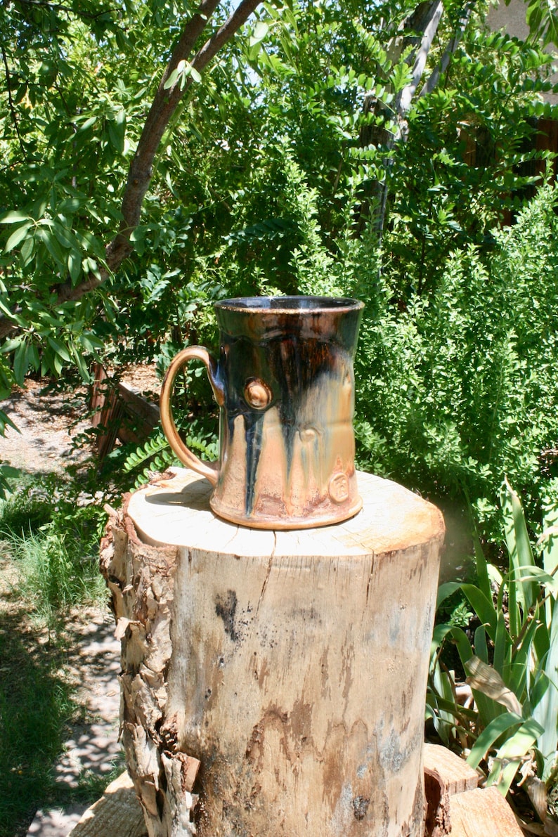 May include: A handmade ceramic mug with a dark brown and copper glaze. The mug has a textured surface and a rounded handle. It is sitting on a tree stump.