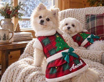 Pode incluir: Dois cães pequenos vestindo vestidos de Natal xadrez vermelho e verde com acabamento em pele sintética branca. Os vestidos apresentam um aplique de árvore de Natal verde, um laço de cetim verde e o nome Anouk bordado em branco na bainha.