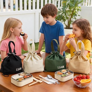 May include: Four fabric drawstring lunch bags in black, cream, dark green, and tan, each featuring a top handle and the name LIAM printed in white. Two rectangular metal bento boxes with divided compartments and wooden chopsticks are placed on the table.