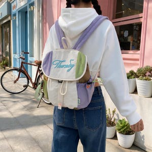 May include: A color-blocked backpack featuring a white central panel, lavender side panels, and a light green accent panel. The flap displays embroidered text in a cursive blue font. It includes lavender shoulder straps, a drawstring closure, and a small tag.