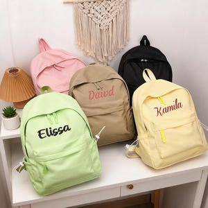 May include: A collection of five backpacks in light green, pink, tan, black, and yellow. The light green bag features the name Elissa, the tan bag features the name Daivd, and the yellow bag features the name Kamila embroidered on the front pocket.