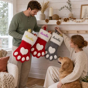 May include: Three Christmas stockings shaped like animal paws with white faux fur cuffs and heart-shaped pads. Two stockings are red with yellow and purple embroidery reading Aurelia, and one is grey with black embroidery reading Aurelia.