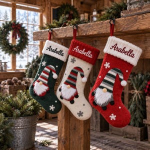 May include: Three knitted Christmas stockings hanging from a wooden mantelpiece. Each stocking features a gnome with a striped hat, a white faux fur beard, and the name Arabella embroidered on the white cuff. The stockings are dark green, cream, and red.