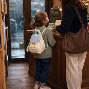 May include: A small canvas backpack featuring a light blue flap, a mint green top handle, and a cream-coloured body. The bag has a drawstring closure, a front pocket with the name Seraphine embroidered in cursive, and adjustable shoulder straps.