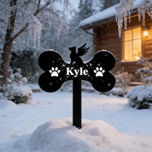 May include: Black metal pet memorial stake in a bone shape, with the name "Kyle" and paw prints. A silhouette of a dog with wings sits atop the bone. The stake is in a snowy outdoor setting.