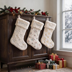 Può includere: Tre calze natalizie in pelliccia sintetica bianca con risvolti in peluche, personalizzate con i nomi ricamati Sophia, Ethan e Mia. Le calze presentano motivi a fiocchi di neve argentati, multicolori e dorati, appese a un mobile in legno scuro.