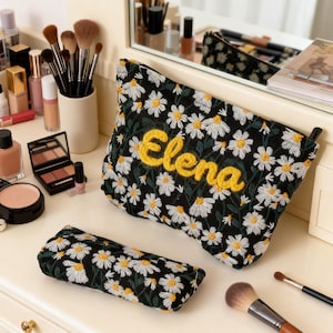 May include: Black cosmetic bag and pencil case embroidered with white daisies. The word "Elena" is embroidered in yellow on the cosmetic bag. Makeup brushes, compacts, and other beauty products are on the vanity.