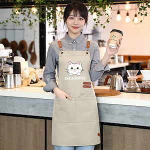 Puede incluir: Delantal beige con un gráfico de gato y el texto "CAT & COFFEE". El delantal tiene correas de cuero marrón y bolsillos. La persona sostiene una taza de café.
