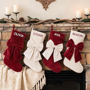 May include: Four velvet Christmas stockings hanging in a row, alternating between deep red and cream colors. Each stocking features a large, matching velvet bow and is personalized with white or dark red embroidered names: Olivia, Ethan, Ava, Mason.