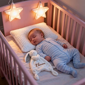 Pode incluir: Um bebê dormindo em um berço rosa, vestindo um macacão listrado azul e branco. Uma almofada de crochê branca e um cobertor de segurança de coelho branco com o nome "Eleven" estão no berço. Duas luzes em forma de estrela pendem acima do berço.
