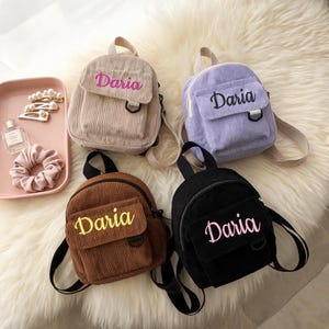 May include: Four small corduroy backpacks in beige, lavender, brown, and black, each featuring the name Daria embroidered on the front pocket. A pink tray sits to the left containing three pearl hair clips, a perfume bottle, and a pink scrunchie.