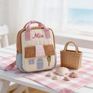 May include: A quilted backpack featuring a house design with the name Mia embroidered in pink. The bag has tan, cream, and light blue panels with pink straps. Beside it sits a small woven basket and several seashells on a checkered surface.