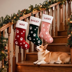 May include: Three Christmas stockings in red, dark green, and light pink, each featuring a white plush cuff with the names MAX, BAILEY, and LUCKY embroidered in red. The stockings are decorated with white paw prints and snowflake patterns.