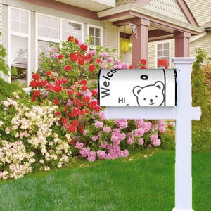May include: White mailbox featuring a black and white bear design, with the words "Welcome" and "Hi". The mailbox is mounted on a white post, set against a backdrop of a house and a garden filled with red, pink, and white flowers.