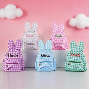 Op de afbeelding: Vijf kleine rugzakken met konijnenoren, elk met een gingham-patroon in paars, grijs, roze, blauw of groen. Elke tas heeft een voorvak met rits en is gepersonaliseerd met geborduurde namen: Daria, Elissa of Kamila in verschillende kleuren.
