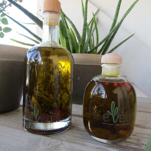 Può includere: Due bottiglie di vetro di olio d'oliva con tappi di legno. La bottiglia più grande contiene erbe, mentre quella più piccola ha grani di pepe rosso. Entrambe le bottiglie hanno il logo "e veá".