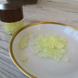 May include: A small glass jar with a wooden lid and a white ceramic plate with a gold rim. Both contain pale yellow, round, resin-like beads. The beads are scattered on the plate and inside the jar.