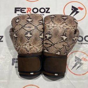 Puede incluir: Un par de guantes de boxeo con un estampado de piel de serpiente en tonos marrones y beige. Las correas de muñeca son de color marrón oscuro liso. Los guantes tienen el nombre de la marca "FEROOZ".