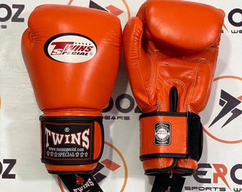 Könnte beinhalten: Paar orangefarbene Boxhandschuhe mit schwarzen Handgelenksriemen. Die Handschuhe tragen das Logo und den Text "Twins Special" am Handgelenk und an der Oberseite. Aus Leder gefertigt, mit einem angebrachten Etikett.