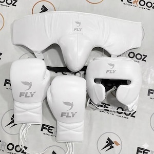 Może przedstawiać: Biały zestaw bokserski, w tym rękawice, ochraniacz głowy i ochraniacz krocza. Każdy element ma napis "FLY" i stylizowane logo ptaka. Zestaw przeznaczony jest do sportu i treningu.