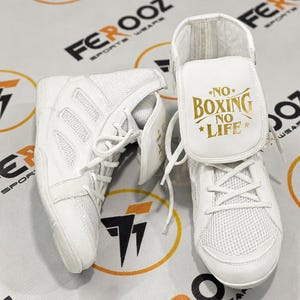 Puede incluir: Par de zapatillas de boxeo blancas con paneles de malla y cuero. Las zapatillas tienen cordones y un diseño de caña alta. La lengüeta de una zapatilla presenta el texto "NO BOXING NO LIFE" en letras doradas. Zapatillas para boxeo y otros deportes de combate.