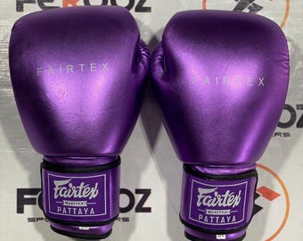 Könnte beinhalten: Paar metallisch-lila Fairtex Muay Thai Boxhandschuhe. Die Handschuhe tragen das Fairtex-Logo und das Wort "Pattaya" auf den Handgelenksriemen. Sie sind für das Training und den Wettkampf in Kampfsportarten konzipiert.