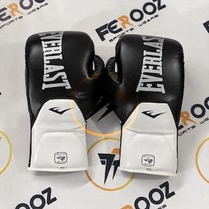 Puede incluir: Un par de guantes de boxeo Everlast en blanco y negro. Los guantes tienen la palabra "EVERLAST" impresa en letras blancas grandes en la parte superior. Los guantes tienen una sección blanca en la palma y las palabras "HECHO EN MEXICO".